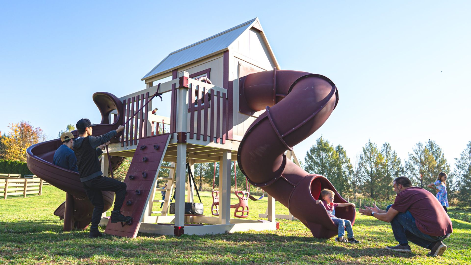 children playing on a large maroon and tan playset with swing set accessories including a climbing wall and two spiral tube slides